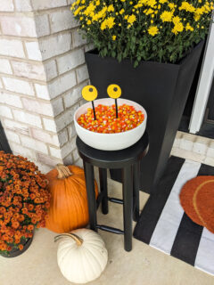 Monster Candy Bowl with candy corn sitting on a stand on a front porch with pumpkins and mums