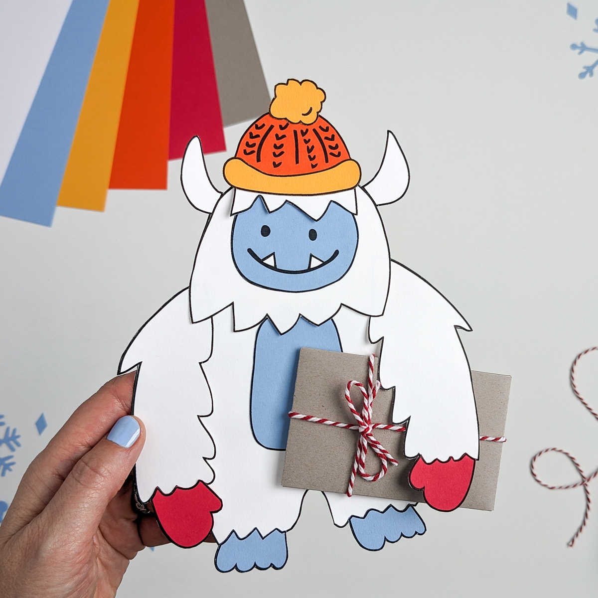 Paper Yeti wearing a winter hat and mittens and carrying a gift card wrapped as a brown paper package tied up with strings