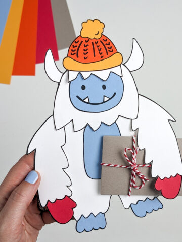 Paper Yeti wearing a winter hat and mittens and carrying a gift card wrapped as a brown paper package tied up with strings